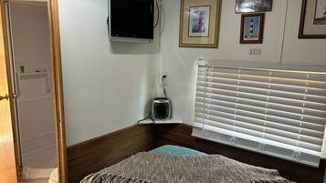 Un dormitorio con cama, televisor montado en la pared, ventana con persianas y un cuadro enmarcado.