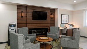 Un espace lounge moderne avec des fauteuils gris, une table basse ronde en bois, un téléviseur à écran plat fixé à un mur en bois, un distributeur automatique de billets et un bureau avec un ordinateur.