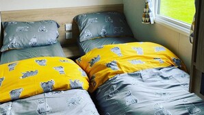 A room with two beds, each with a patterned duvet and pillows, a window with curtains, and a view of a grassy yard outside. - Charming 3-Bed Holiday home at Primrose Valley (Filey)