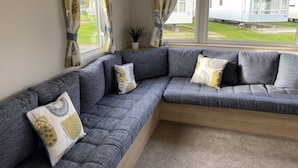A modern living room with a grey sectional sofa, patterned cushions, and a view of a residential area through large windows. - Charming 3-Bed Holiday home at Primrose Valley (Filey)