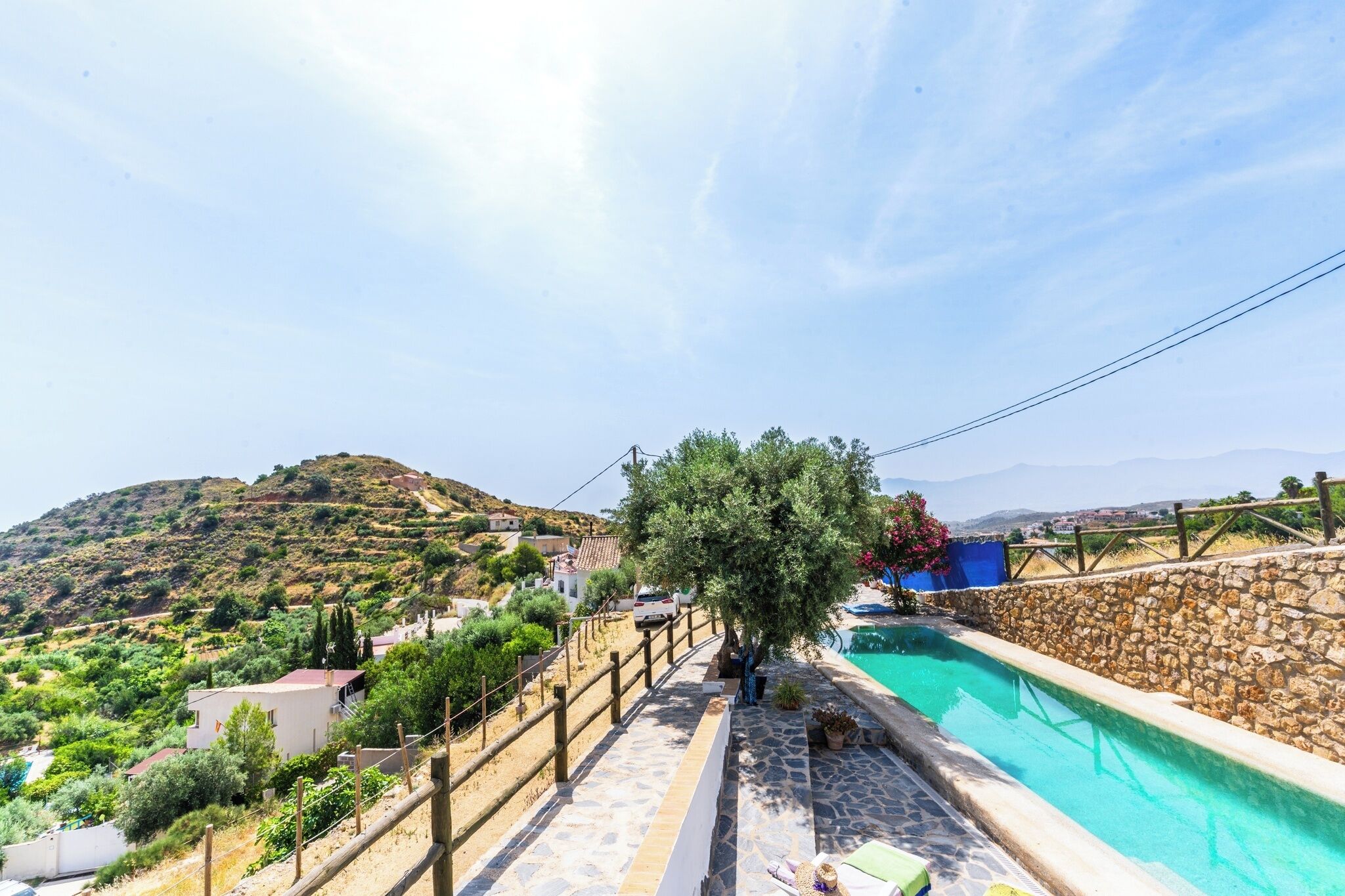 A pool area with stone flooring, a wooden fence, and a view of a hillside with houses.