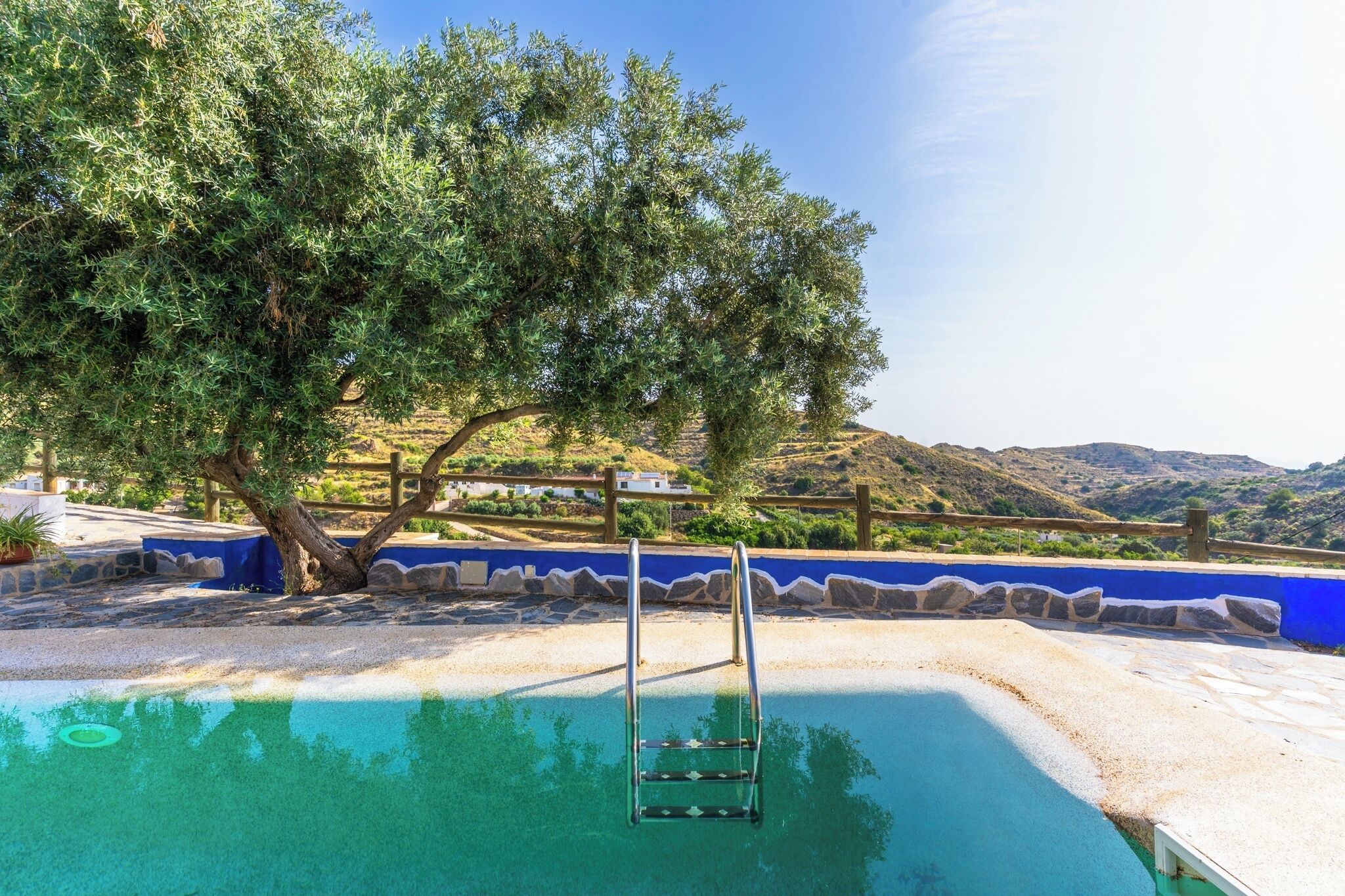 Una piscina con escalera metálica, rodeada por un suelo de baldosas azules y blancas, con un árbol y vistas a las colinas al fondo.