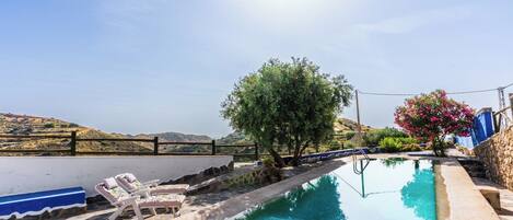 A swimming pool with lounge chairs and a tree, surrounded by stone walls and a view of hills.