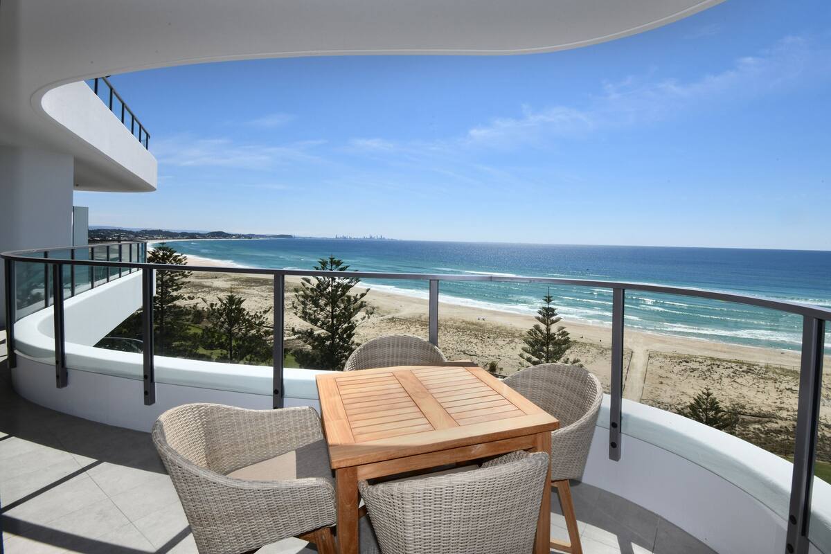 A balcony with a wooden table and wicker chairs overlooking a beach.