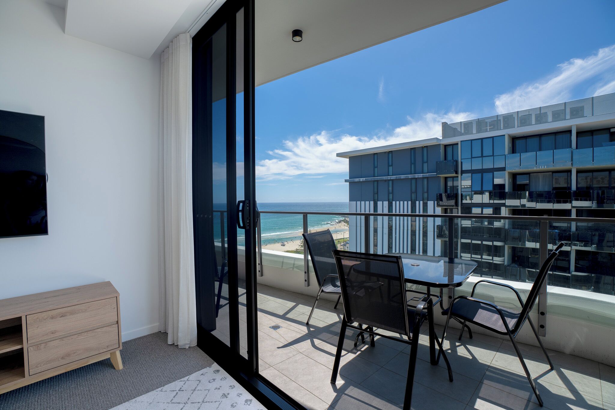 A balcony with a glass door, a table with chairs, and a view of the beach and ocean.