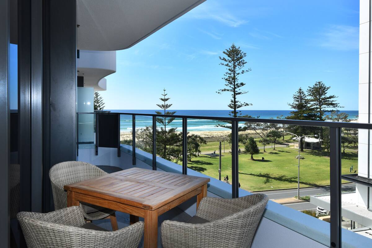 A balcony with wicker furniture, a wooden table, and a view of the beach and ocean.