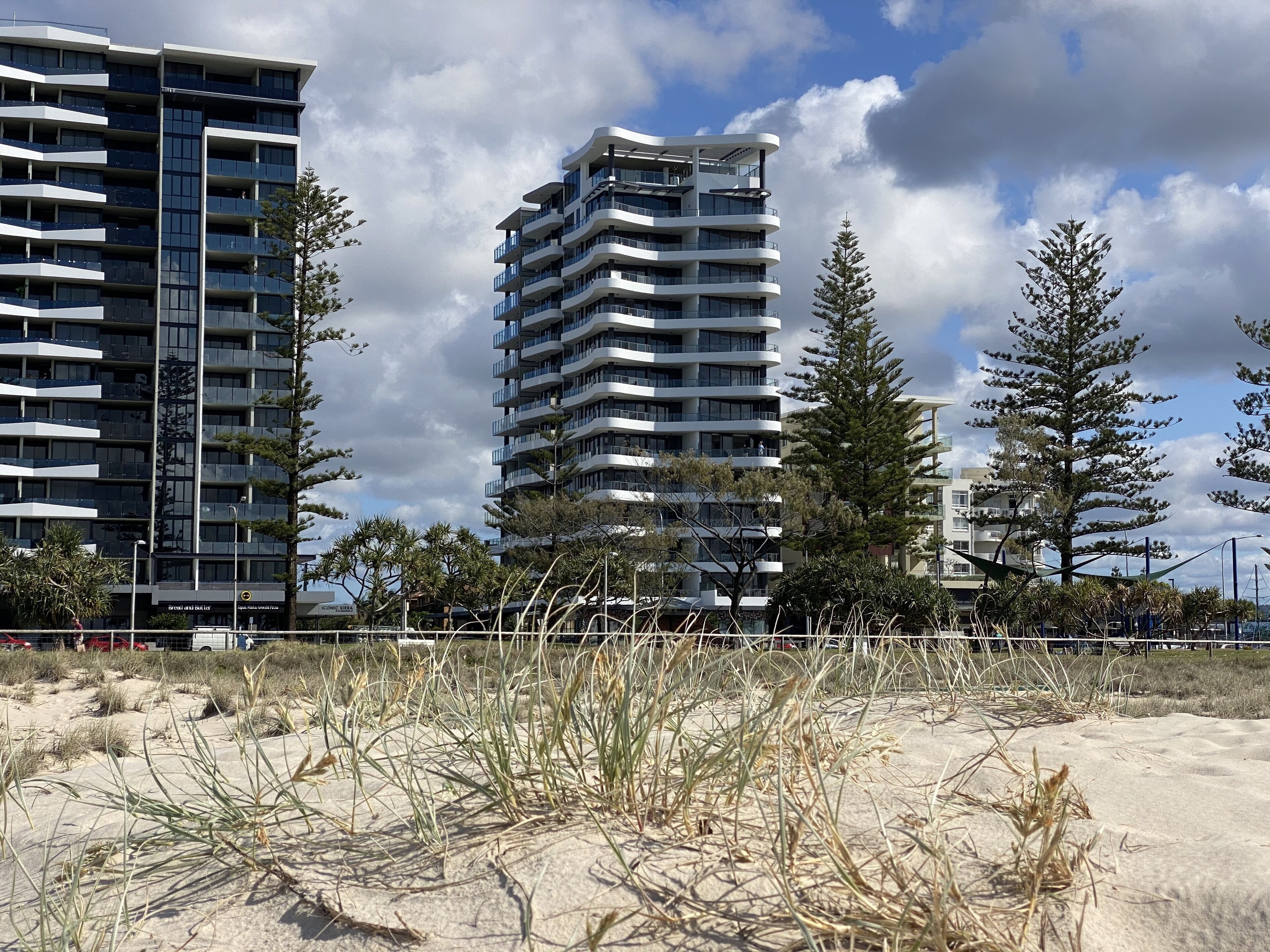 A modern high-rise building with multiple balconies and a sandy foreground with sparse vegetation.