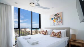 A hotel room with a large bed, a ceiling fan, a painting, a nightstand, and a view of the beach. - X Kirra Apartments (Coolangatta)