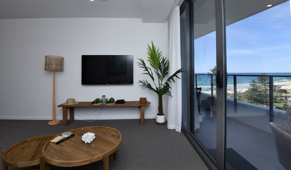 A modern living room with a flat-screen TV, a wooden coffee table, a plant, and a balcony with a view of the beach. - X Kirra Apartments (Coolangatta)