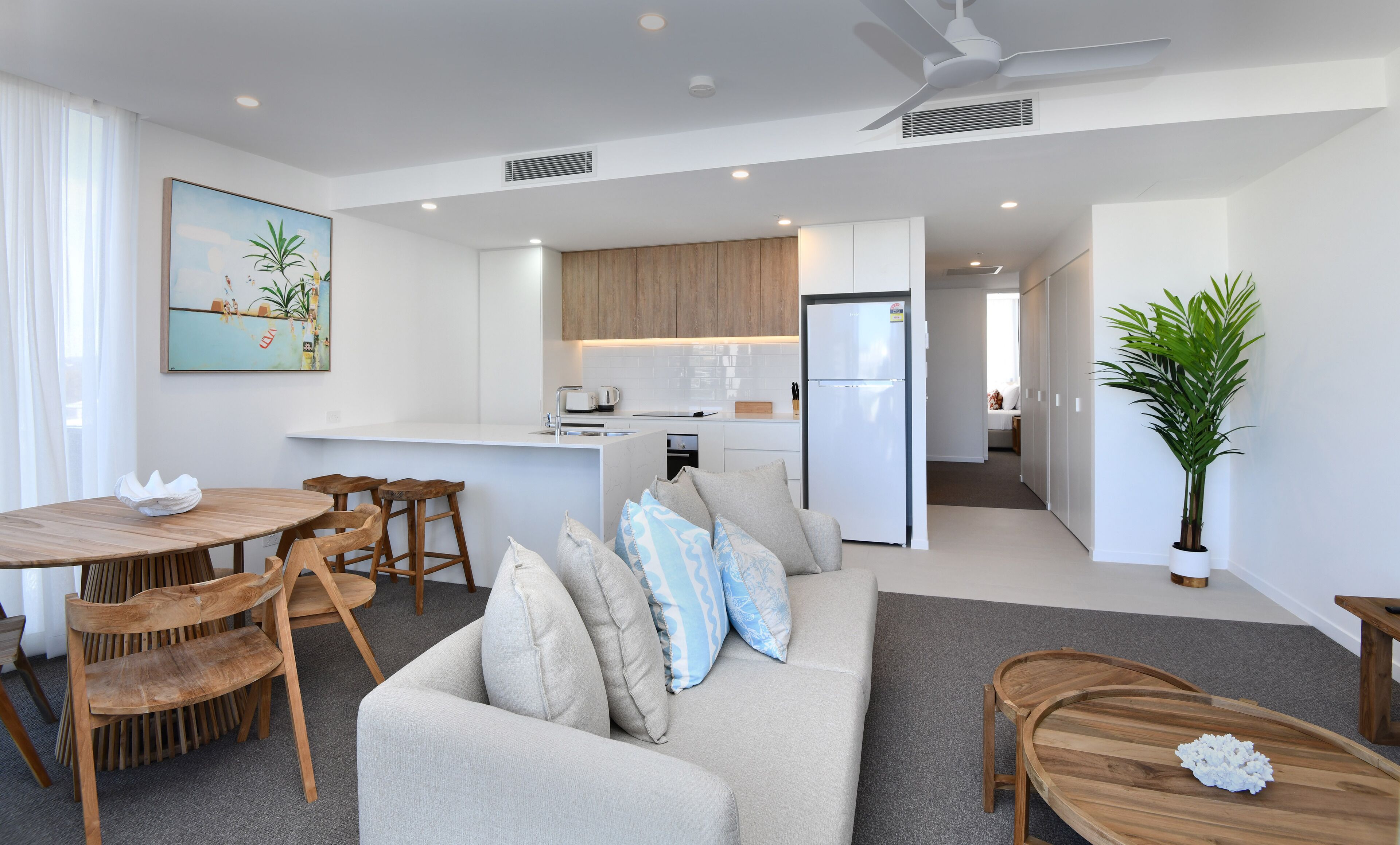 A modern living room with a sofa, wooden chairs, a dining table, and a kitchen area with wooden cabinets and a refrigerator.