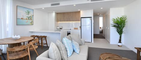 A modern living room with a sofa, wooden chairs, a dining table, and a kitchen area with wooden cabinets and a refrigerator.
