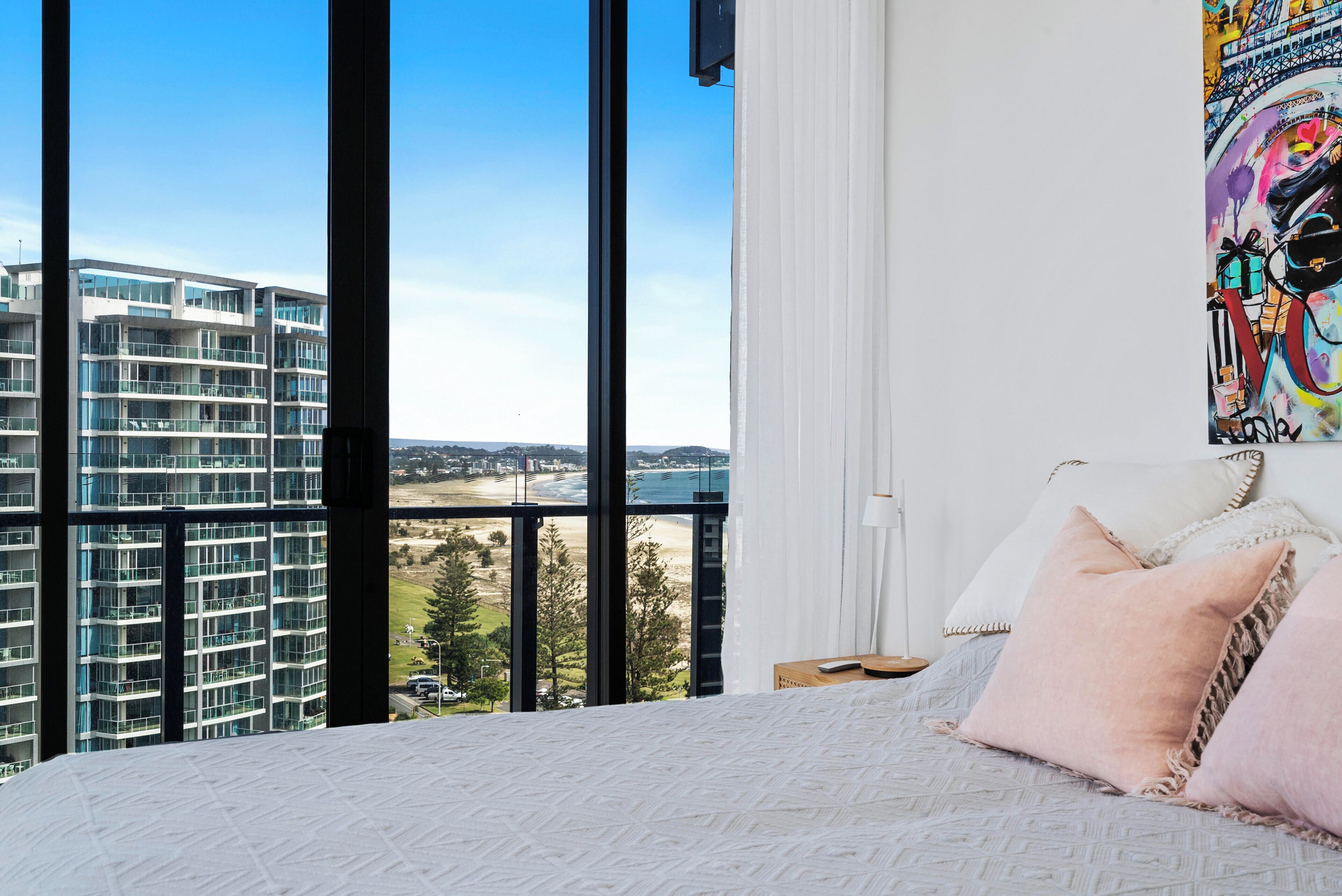 A bedroom with a large window overlooking a modern building and a beach.