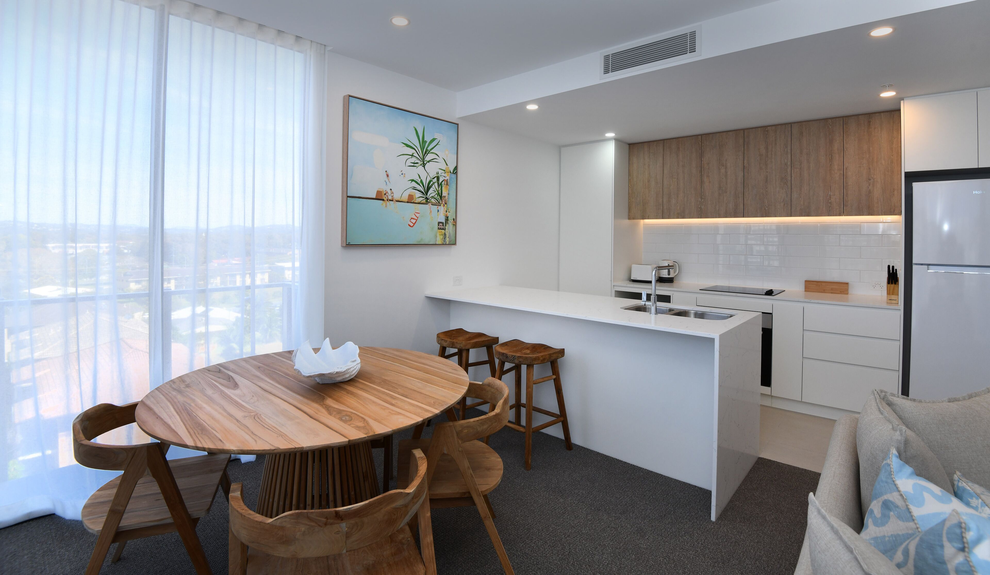 Una cocina moderna con encimera blanca, armarios de madera y un refrigerador empotrado. Hay una zona de comedor con mesa y sillas de madera.