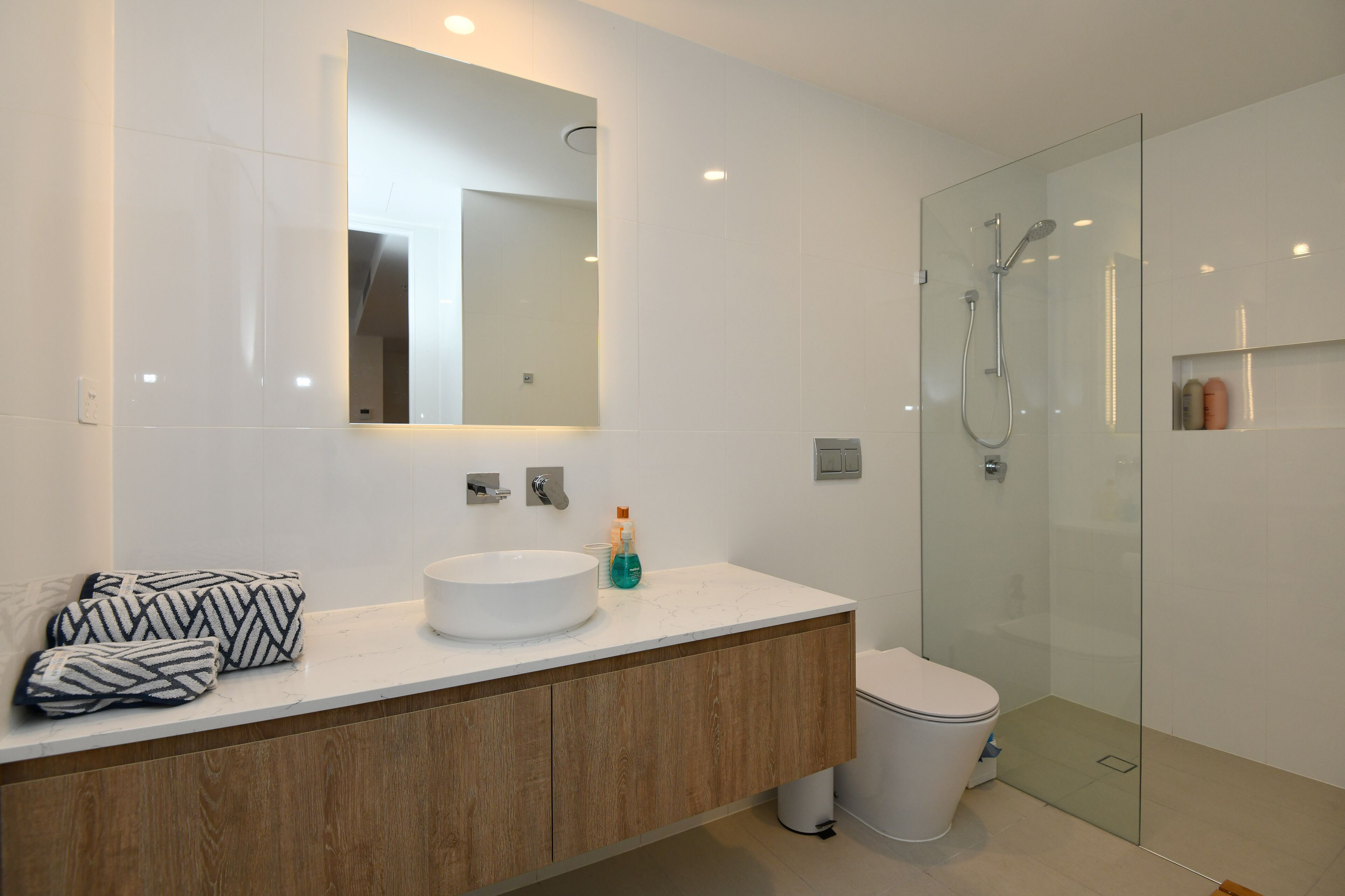 Ein modernes Badezimmer mit einem weißen Waschbecken, einem Holz-Waschtisch, einem großen Spiegel und einer gläsernen Duschkabine.