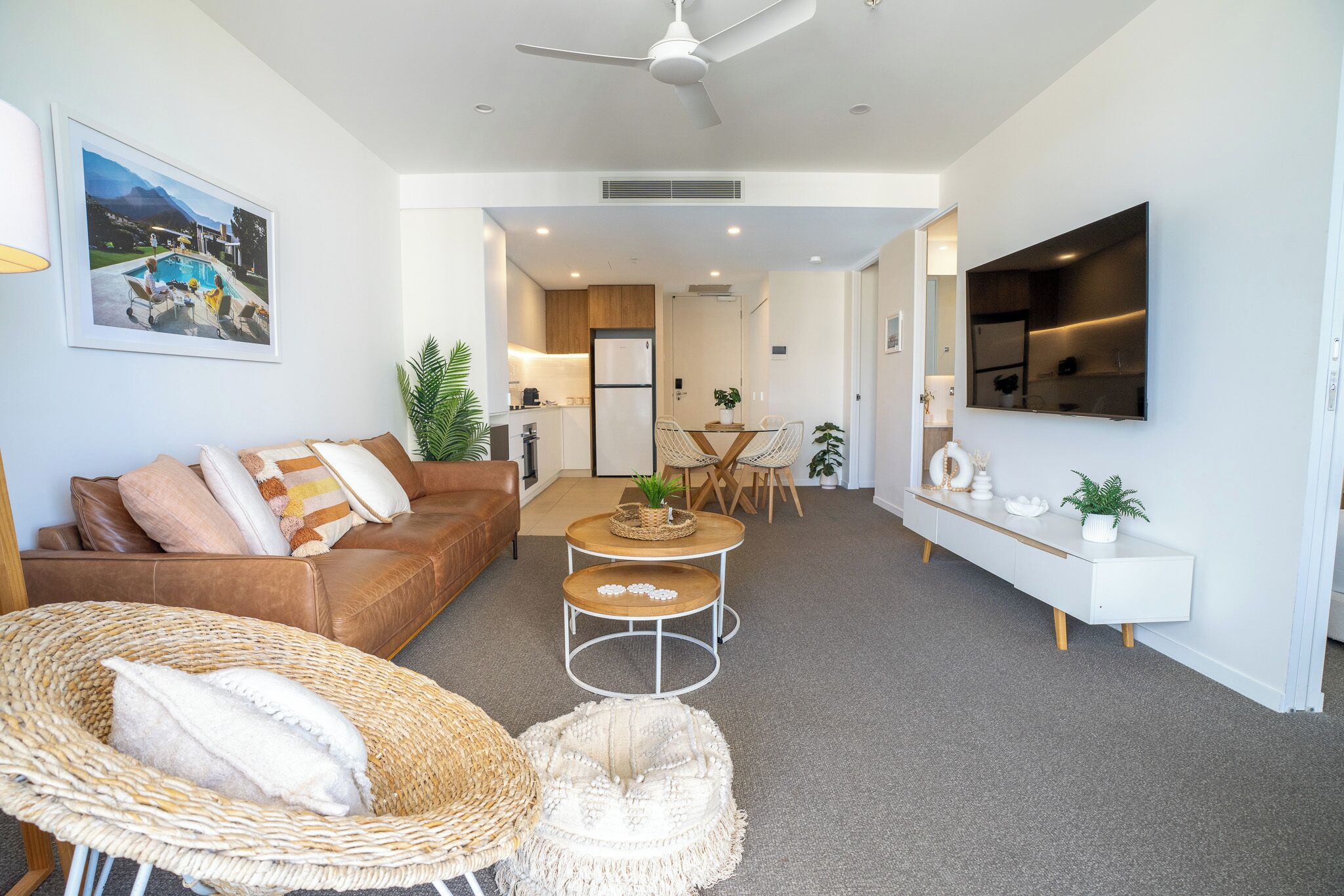 A modern living room with a brown leather sofa, wicker chairs, a small dining table, and a white console table with a TV.