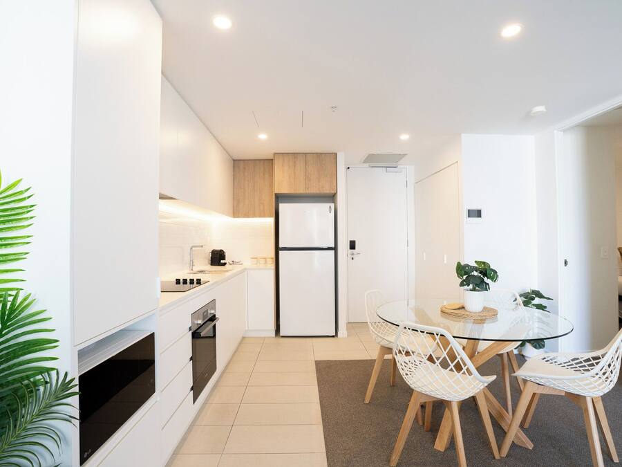 Una cocina moderna con armarios blancos, horno empotrado y refrigerador. Hay un comedor con mesa de vidrio y sillas blancas.