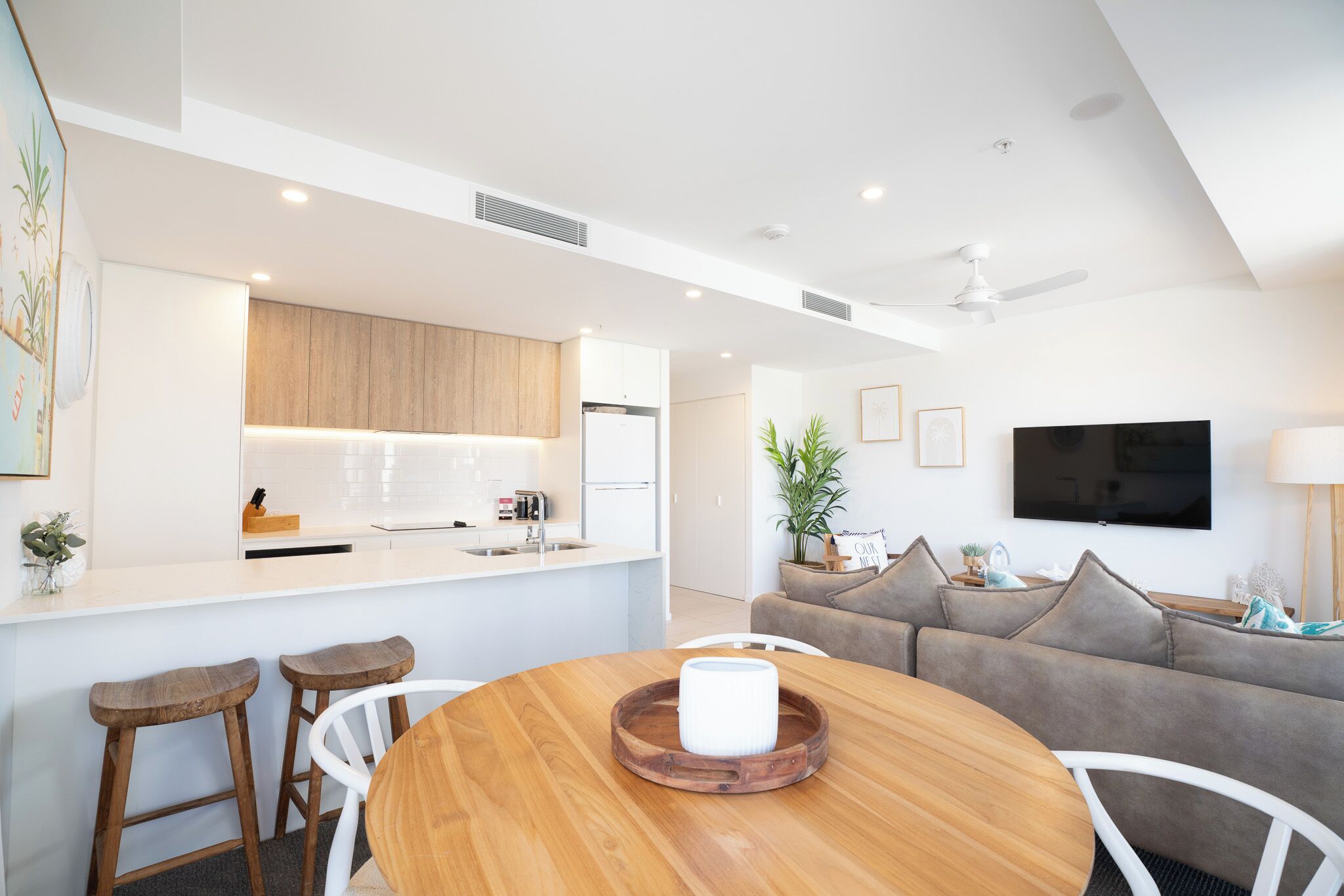 A modern kitchen with white cabinets, a built-in oven, and a refrigerator. There is a dining area with a wooden table and two bar stools. A gray sofa and a TV are visible in the living area.