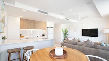 A modern kitchen with white cabinets, a built-in oven, and a refrigerator. There is a dining area with a wooden table and two bar stools. A gray sofa and a TV are visible in the living area.