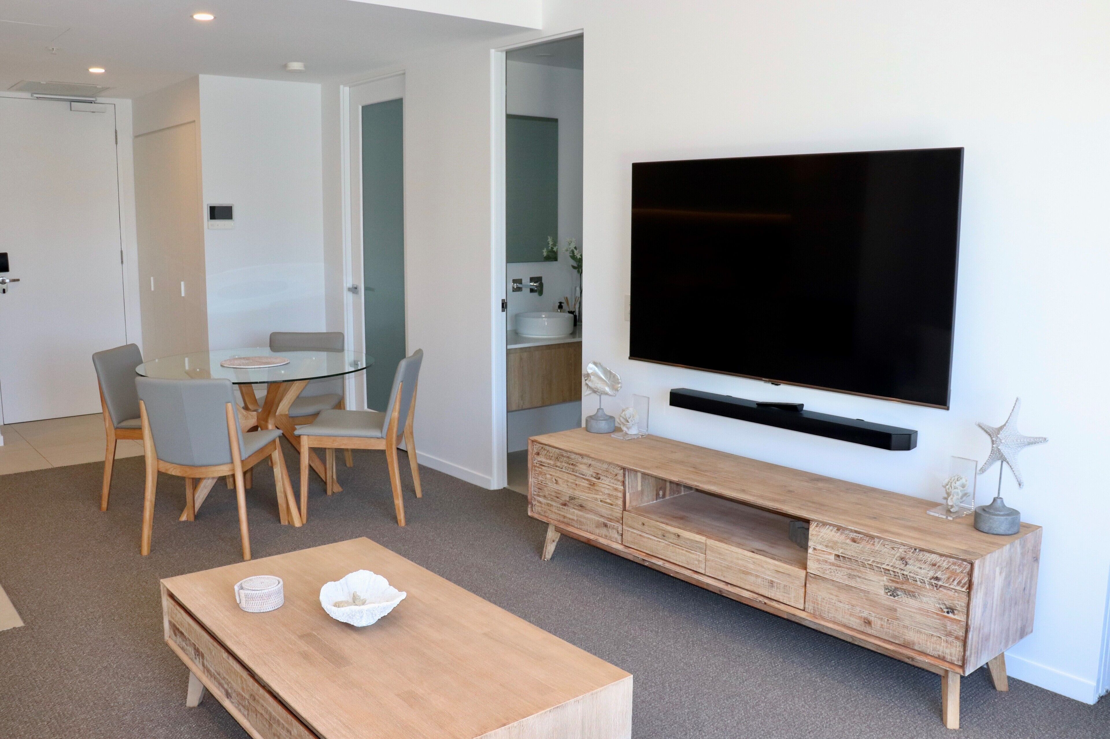 Ein modernes Wohnzimmer mit einem an der Wand befestigten Flachbildfernseher, einem Holztisch und einem Essbereich mit Stühlen und einem Glastisch.