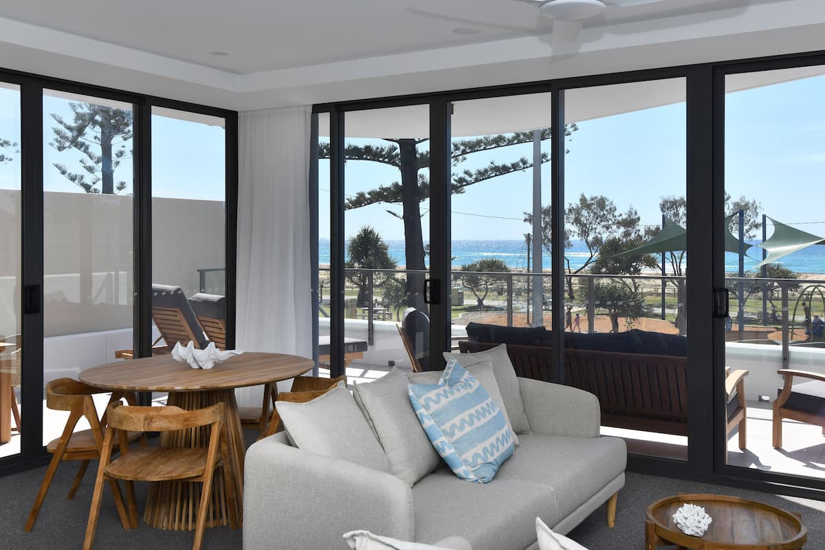 A modern living room with large windows, a round wooden table, and a grey sofa with a patterned pillow.