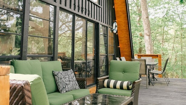 A modern outdoor seating area with wicker furniture and a glass table, overlooking a forested area.