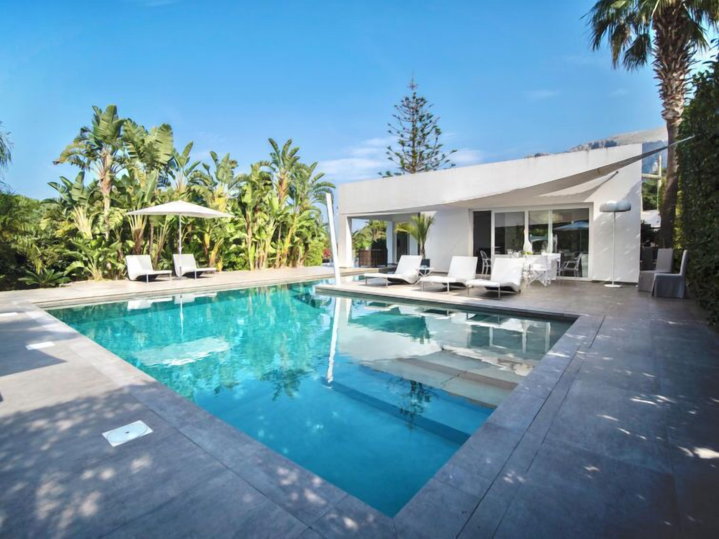 A modern pool area with clear blue water, white lounge chairs, and lush tropical plants.