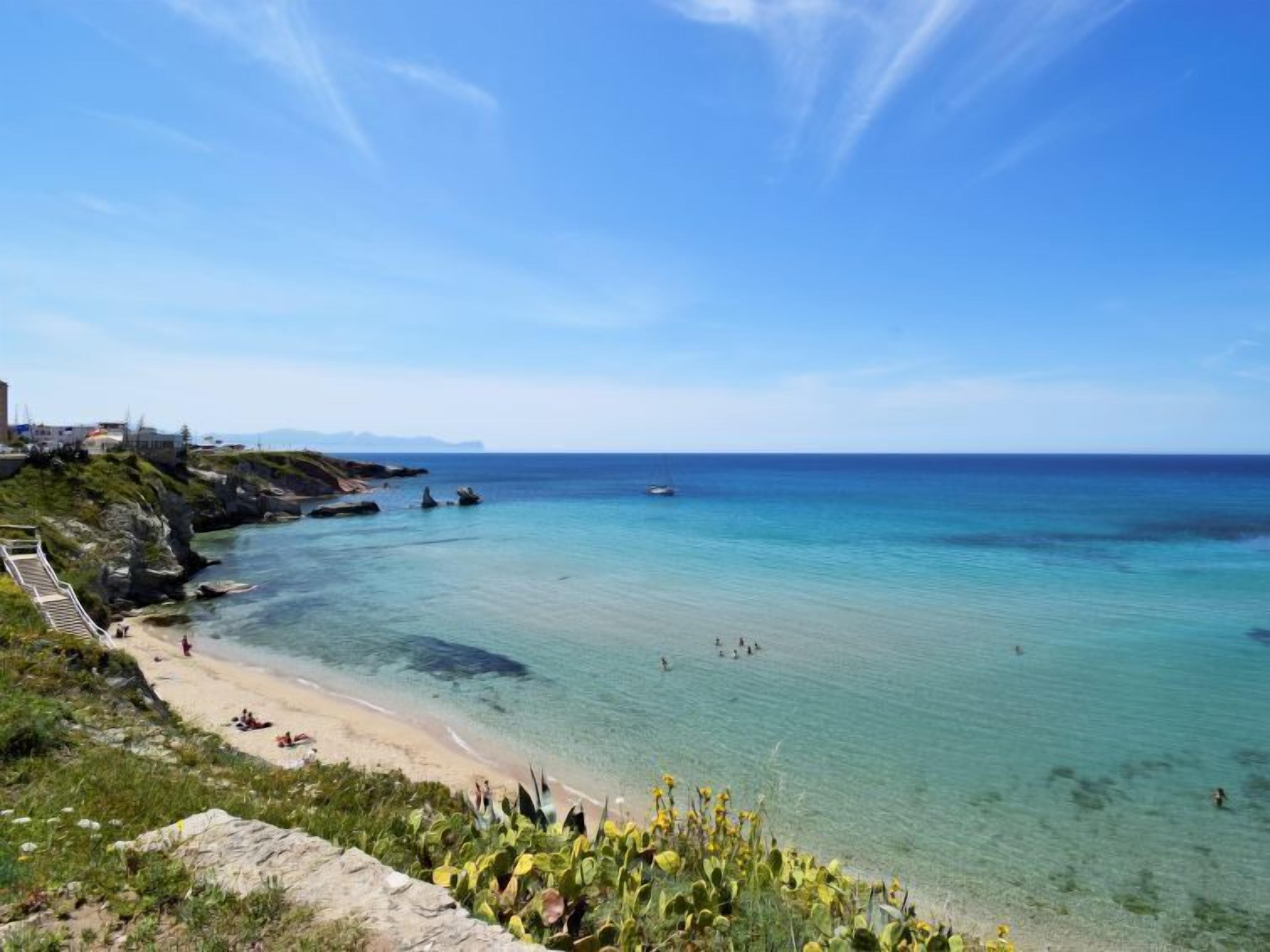 Uma praia costeira com águas cristalinas, areal arenoso e um afloramento rochoso com vegetação.