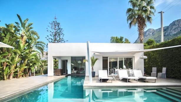 Uma área moderna à beira da piscina, com piscina transparente, espreguiçadeiras brancas e um alpendre envidraçado com vista para as montanhas.