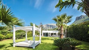 Um gazebo branco num jardim exuberante com piscina e uma casa moderna ao fundo.