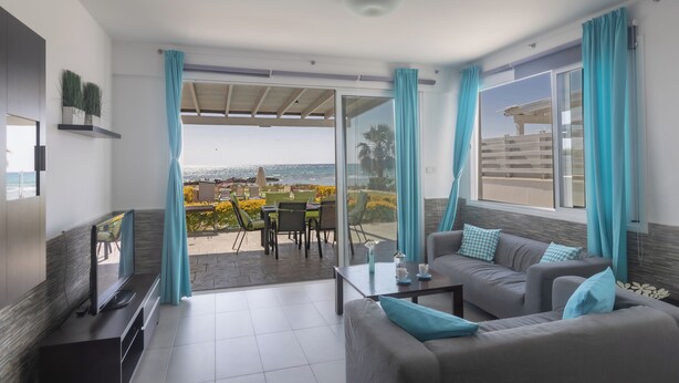 A modern living room with a sofa, TV, and a balcony with outdoor furniture. - Villa Suri, Luxury Seafront Villa (Sotira)