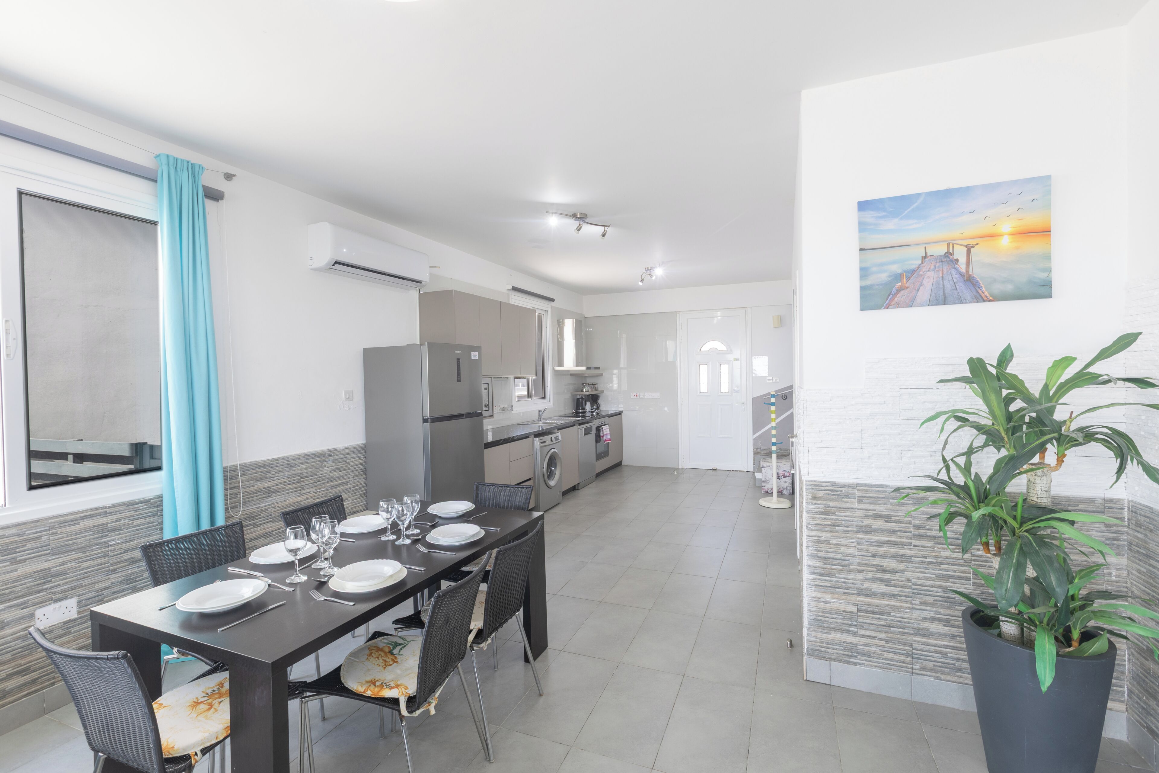A modern kitchen with a dining area, a large potted plant, and a sunset painting on the wall.