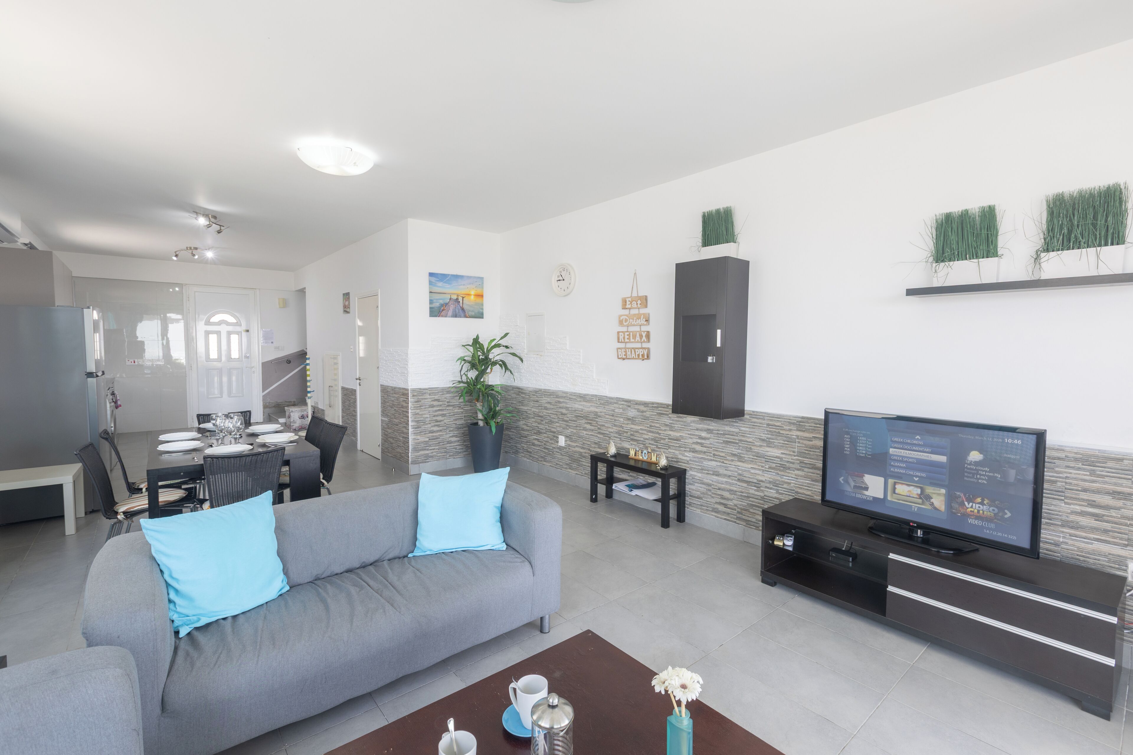 Ein modernes Wohnzimmer mit grauem Sofa, Couchtisch, Fernseher auf einem schwarzen Schrank, Essbereich mit Tisch und Stühlen sowie einer Treppe zu einem weiteren Raum.
