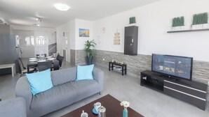 A modern living room with a grey sofa, a coffee table, a TV on a black stand, a dining area with a table and chairs, and a staircase leading to another room. - Villa Suri, Luxury Seafront Villa (Sotira)