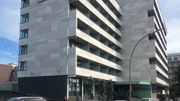 Un hôtel à plusieurs étages, doté d’un logo « AH » bien visible et d’une enseigne sur laquelle on peut lire « un chez-vous ». Il présente un design moderne, avec un toit plat et de grandes fenêtres.
