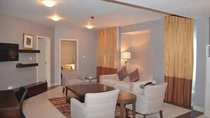 A hotel room with a living area, a flat-screen TV mounted on the wall, a sofa set, a coffee table, and a lamp.