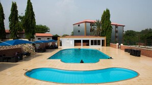 An outdoor swimming pool with a tiled deck, a covered seating area, and a building with a red roof in the background. - Room-grand Ibro Hotel - Guest Room (Abuja)