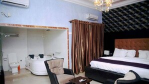 A hotel room with a bed, a sofa, a chair, a small table, a bathtub, a mirror, curtains, and an air conditioning unit. - Room-hampton Towers & Spa - Guest Room (Asaba)