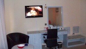 A hotel room with a television mounted on the wall, a small round table with a bowl, a chair, a vanity with a mirror, and a sink. - Room-hotel De Bently - Guest Room (Abuja)