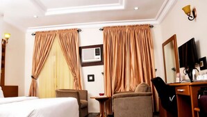 A hotel room with a bed, two armchairs, a desk, a television, and curtains. - Room-hotel De Bently - Guest Room (Abuja)