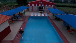 A rectangular pool with a red border, surrounded by lounge chairs and umbrellas, with a building in the background. - Room-hotel De Bently - Guest Room (Abuja)