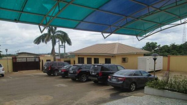 A parking area with several cars, a covered structure, and a building with a yellow facade. - Room-joy House Hotel - Guest Room (Abuja)
