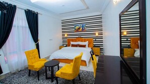 Une chambre d’hôtel avec un lit, deux chaises jaunes, une petite table, un miroir et un tableau accroché au mur.