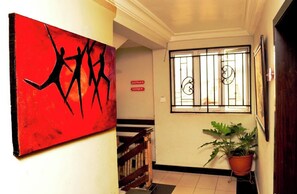 A hallway with a red abstract painting, a staircase, a potted plant, a window with bars, and a sign indicating the lounge area. - Room-prince Of Anthony 1960 Hotel (Lagos)