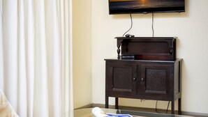 Une pièce dotée d’un téléviseur à écran plat fixé au mur, d’une table basse en verre sur laquelle repose un magazine, d’un meuble en bois foncé et d’un rideau blanc.