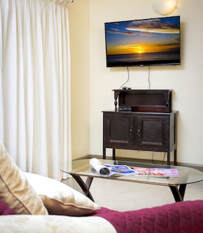 A room with a television mounted on the wall, a glass-top coffee table with a magazine, a dark wooden cabinet, and a bed with a white and pink blanket. - Golden View #115 - by Zenbreak (Holetown)