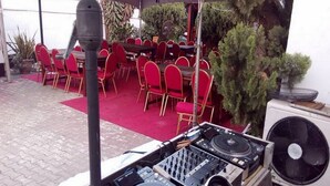 Un área de comedor al aire libre con sillas rojas, equipo de DJ y un aire acondicionado.