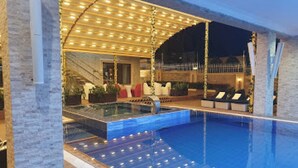 A swimming pool with a sunken seating area, a covered patio with lights, and a view of the surrounding landscape. - Room-the Wells Carlton - Guest Suite (Abuja)
