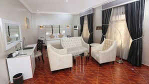 A hotel room with a large bed, two white armchairs, a small table, a mirror, and a large window with curtains. - Room-the Wells Carlton - Guest Suite (Abuja)