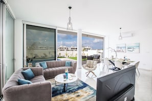 A modern living room with a sofa, coffee table, and TV, a dining area with chairs and a table, and large windows with a view of buildings and sky. - Villa Prol26, Stunning 3bdr Protaras Villa With Pool, Close to Fig Tree Bay (Paralimni)