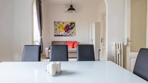 Un comedor con una mesa blanca, cuatro sillas, un cuadro en la pared, un sofá con cojines y un radiador.